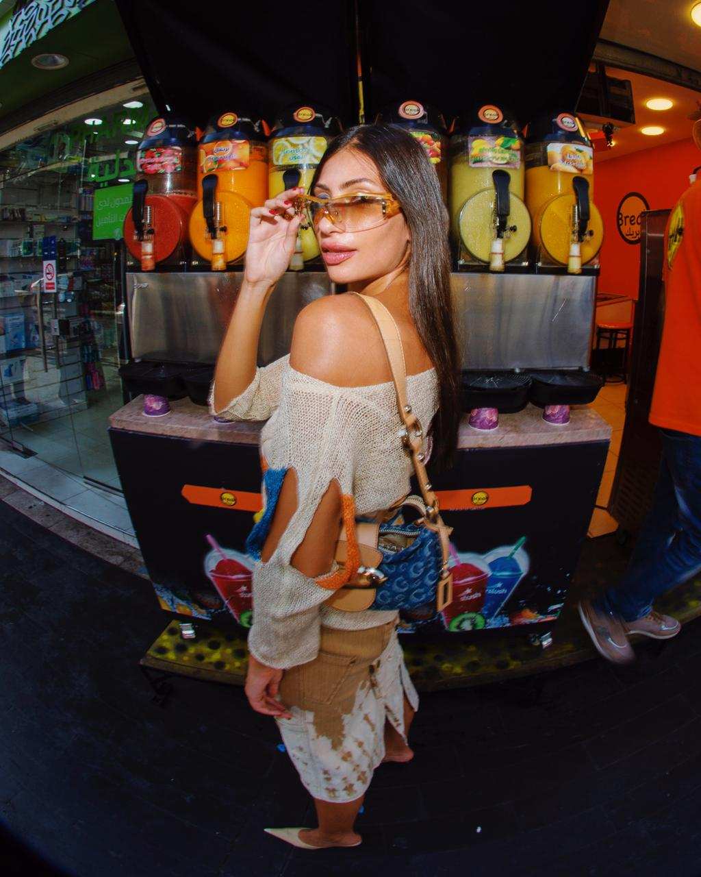 Stylish woman in sunglasses and cutout sweater posing by colorful juice dispensers in urban juice bar