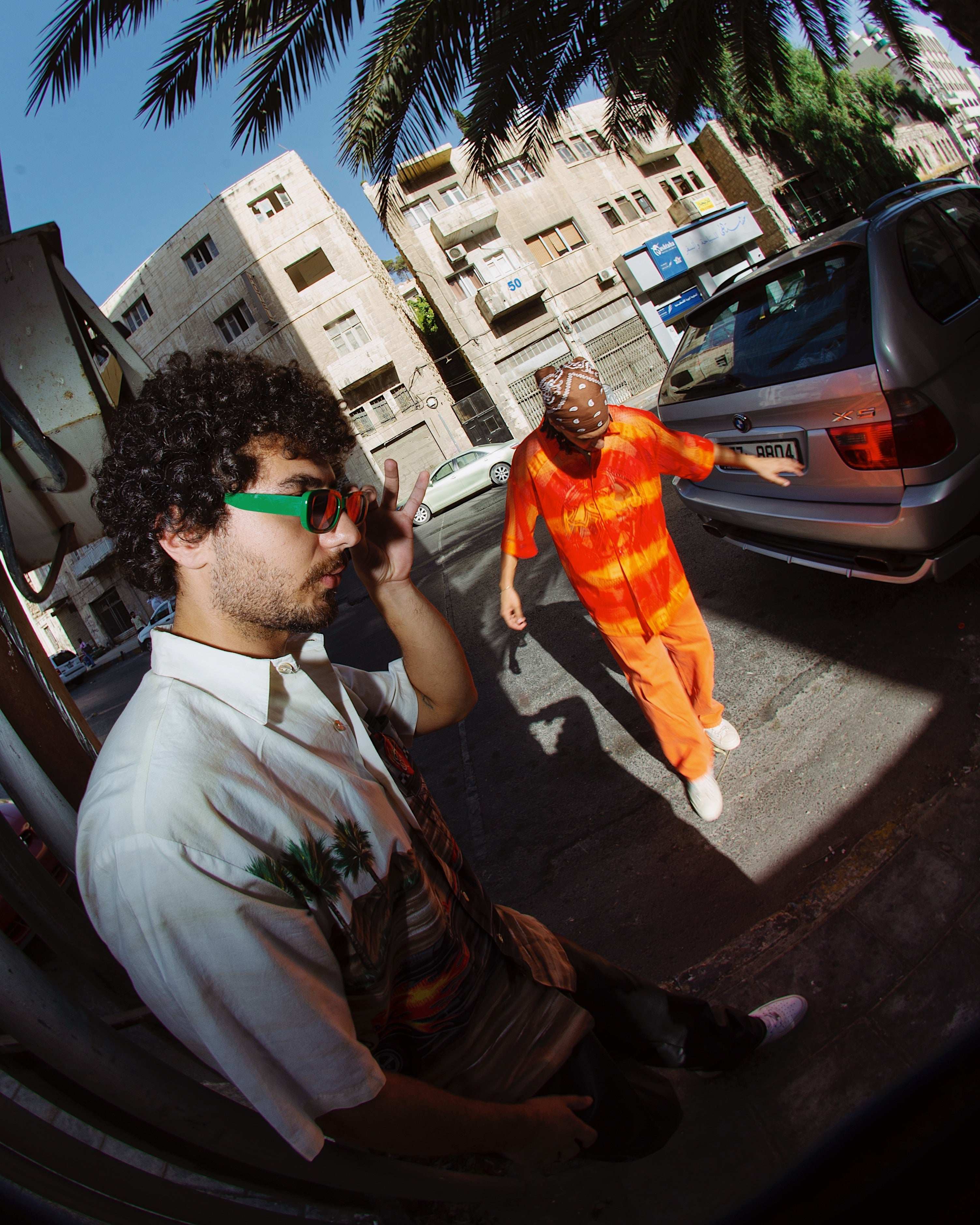 Two men in vintage 90s Y2K streetwear, one in palm print shirt and green sunglasses, other in orange tie-dye jumpsuit, urban palm tree backdrop