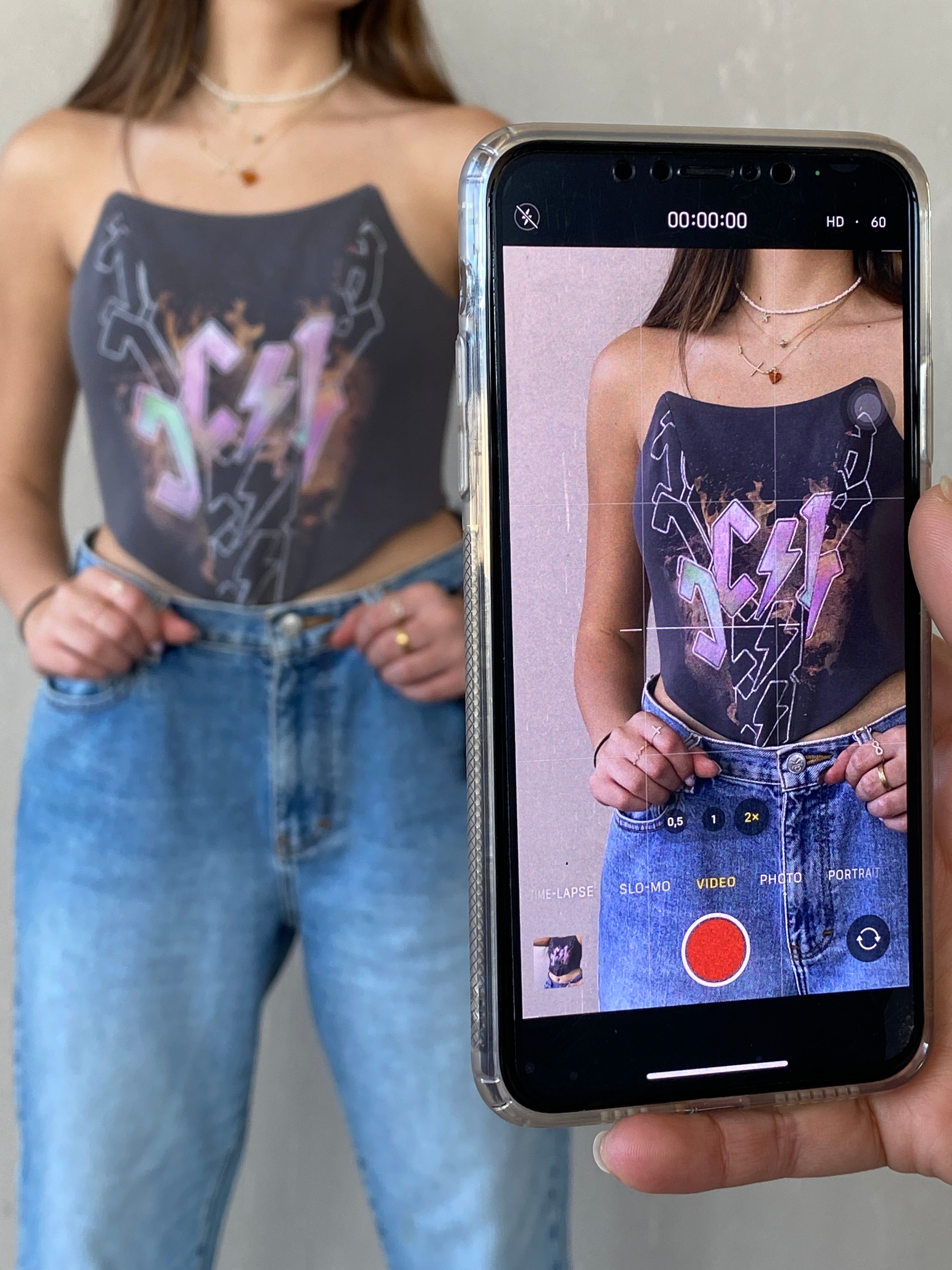 Woman wearing black graphic top and blue high-waisted jeans, captured on phone screen in video mode