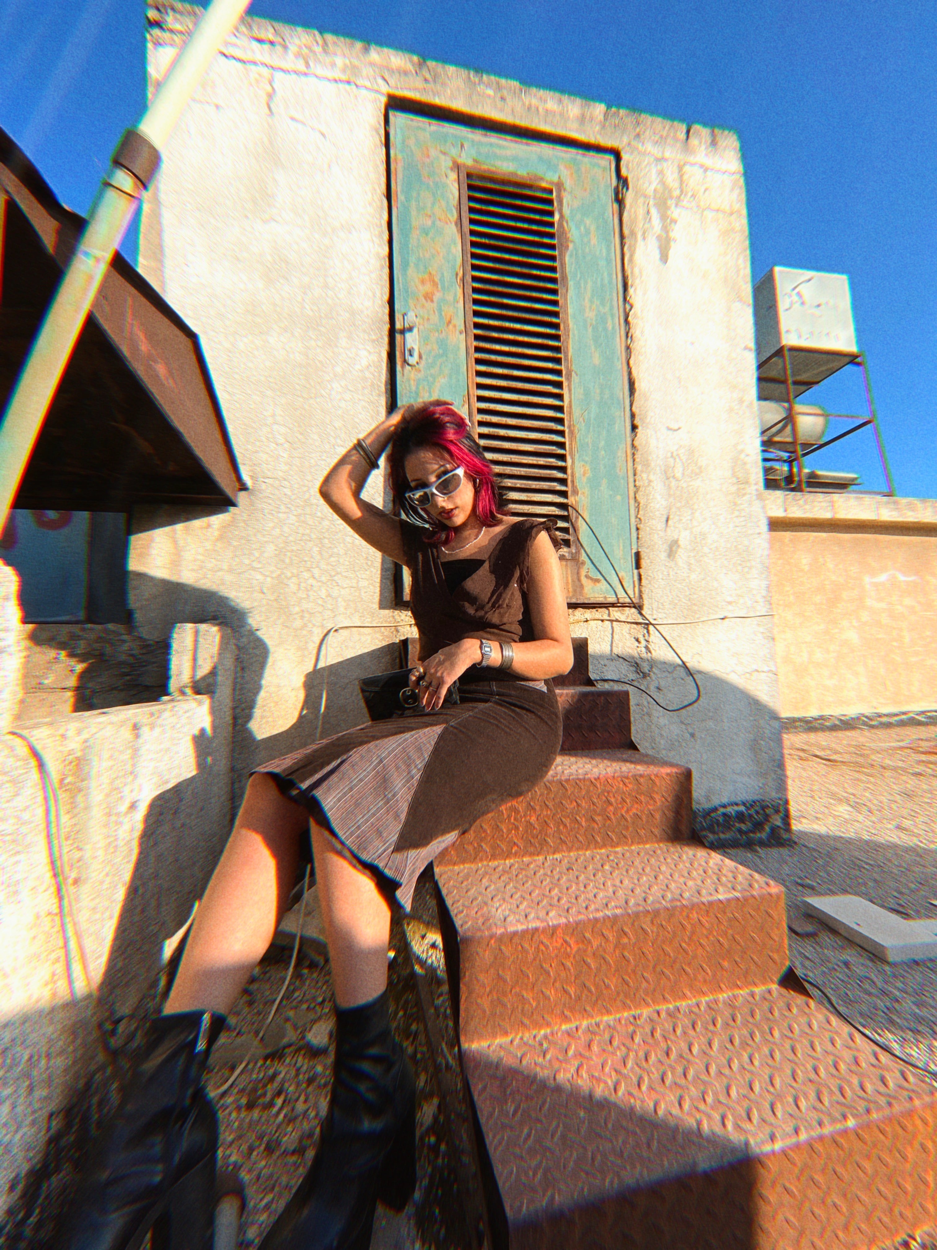 Woman in vintage brown dress & sunglasses sitting on rooftop steps, 90s Y2K fashion vibe