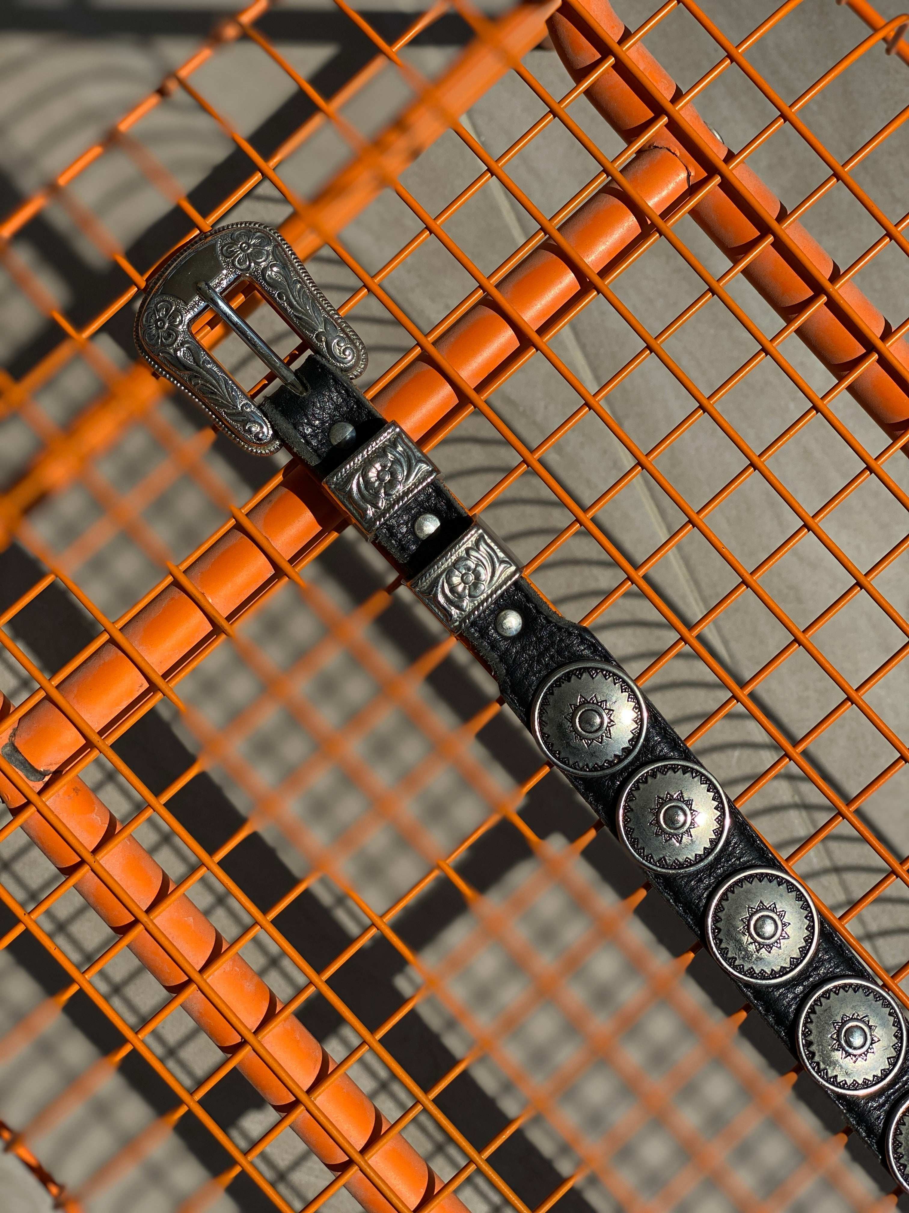 Black vintage leather belt with ornate silver buckle and conchos, styled on orange grid background.