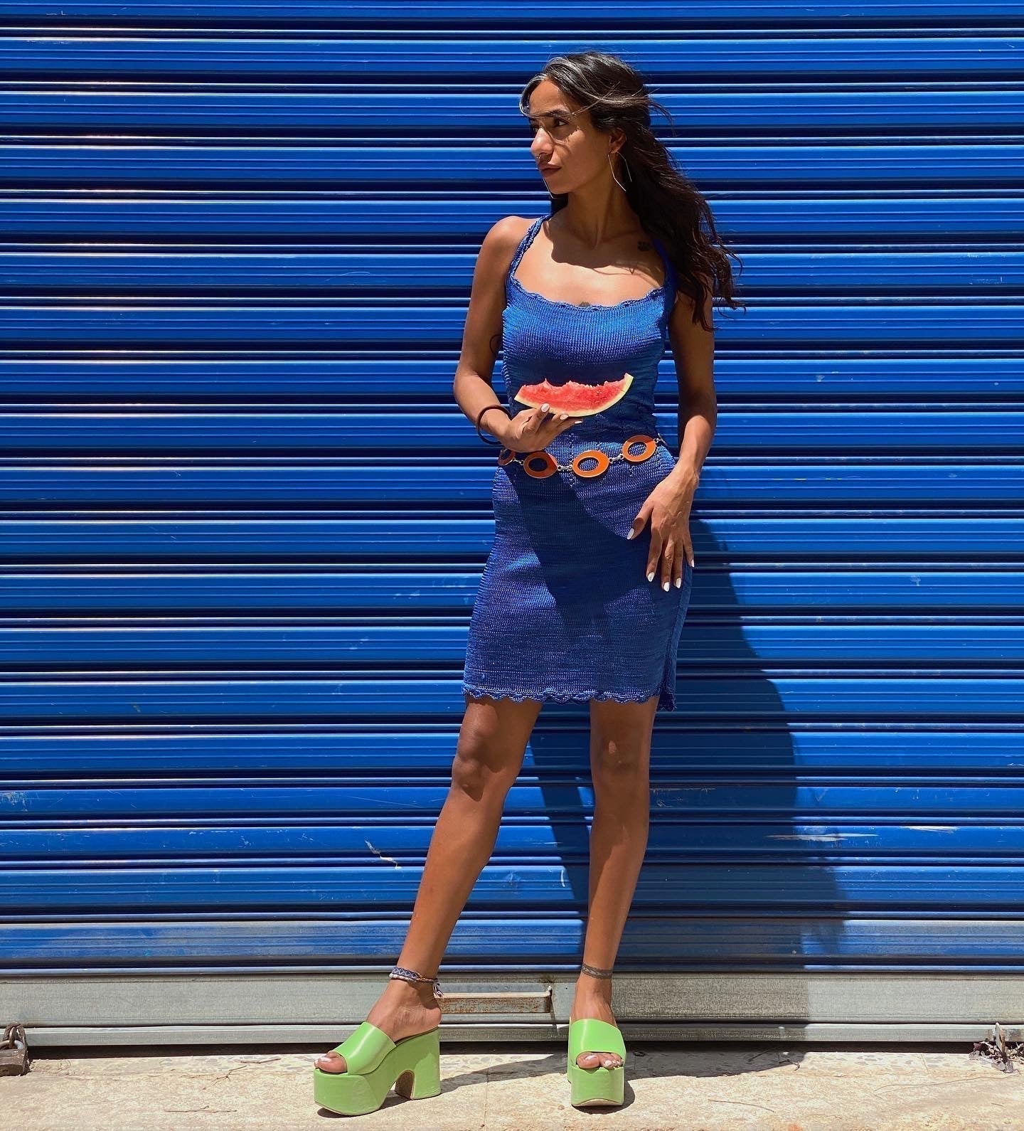 Woman in blue vintage dress with watermelon print, green platform sandals, orange belt, against blue shutter