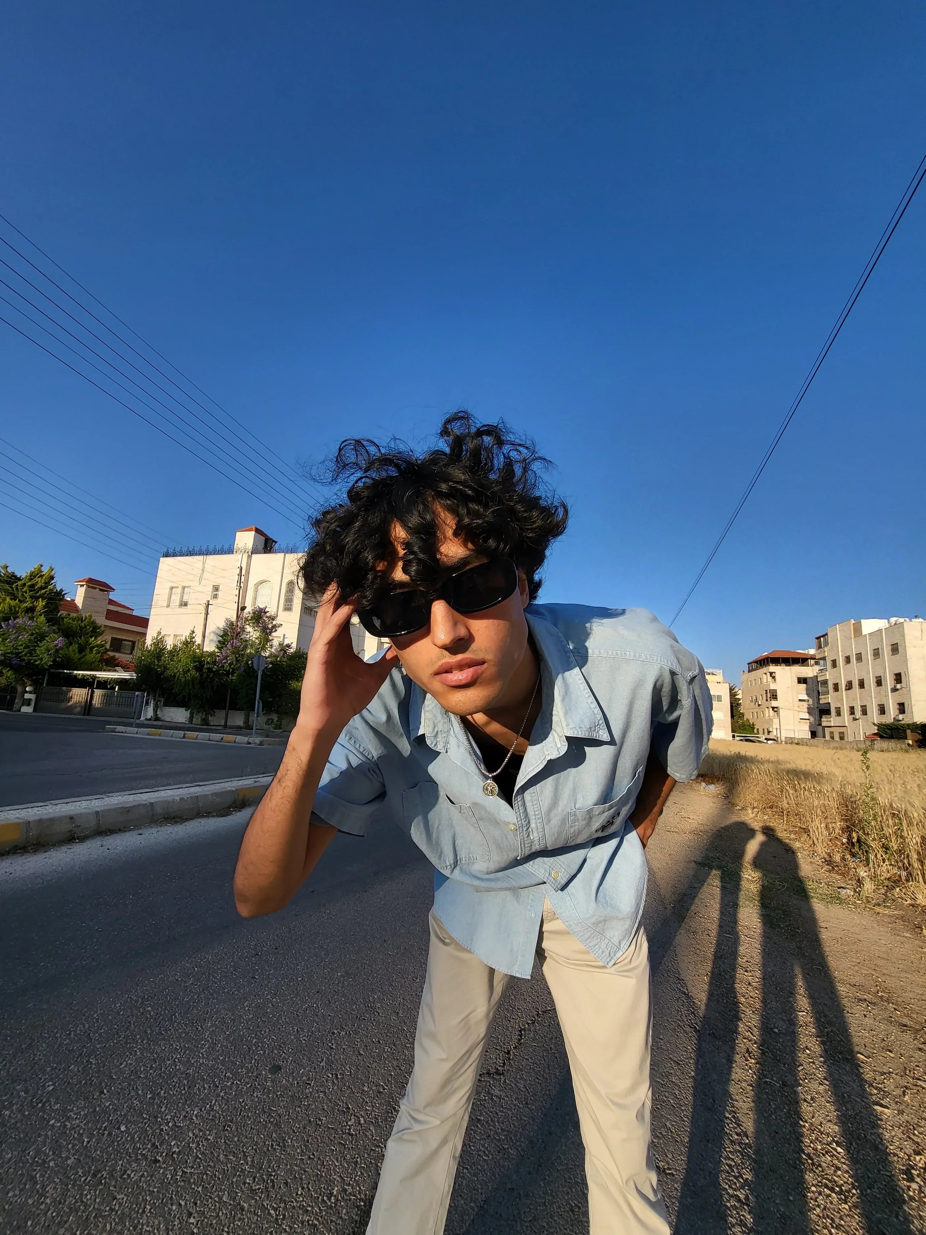 Man in vintage light blue shirt and beige pants, wearing sunglasses, posing on a sunny street