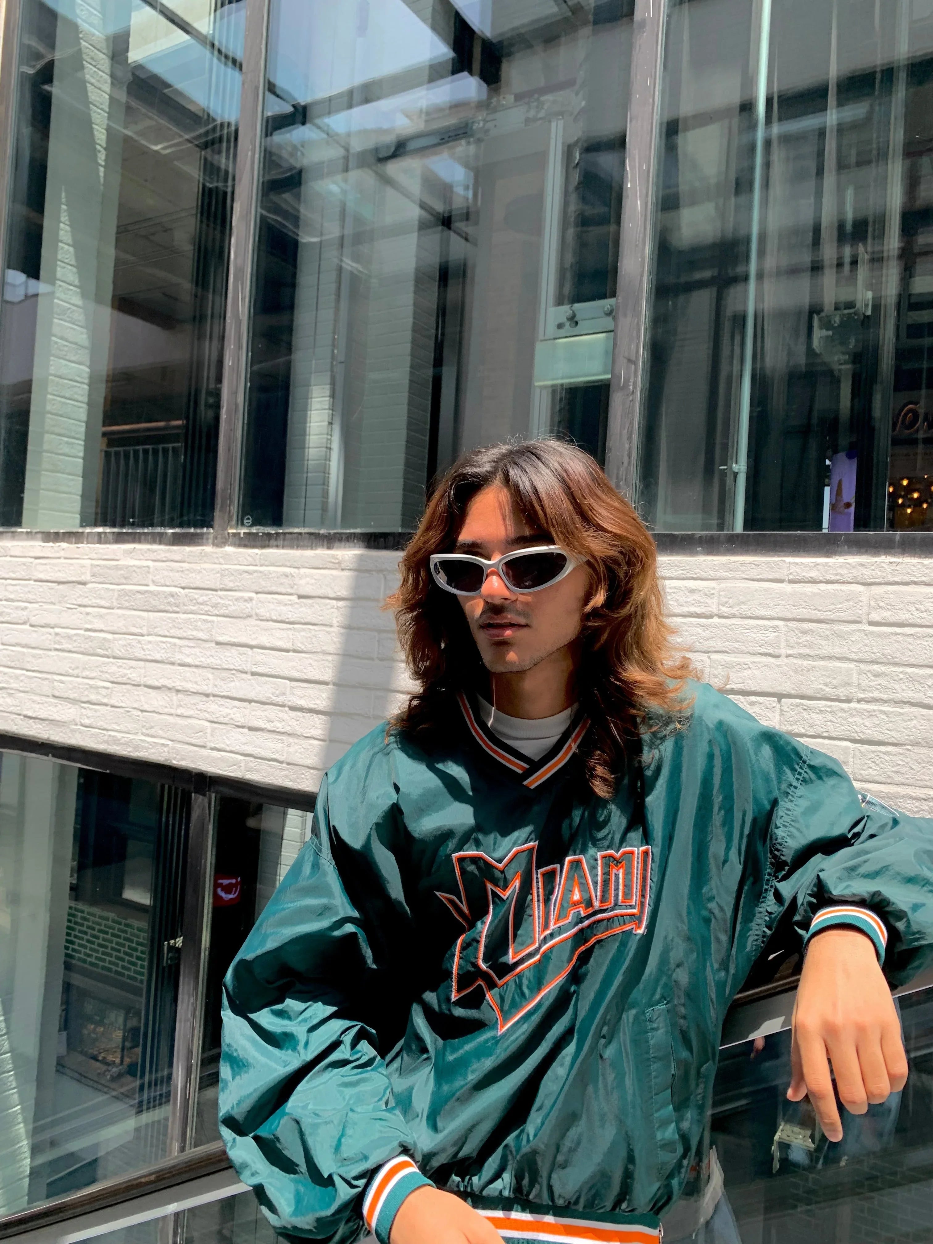 Young man with long hair wearing white cat-eye sunglasses and green Miami sports jacket leaning on glass railing