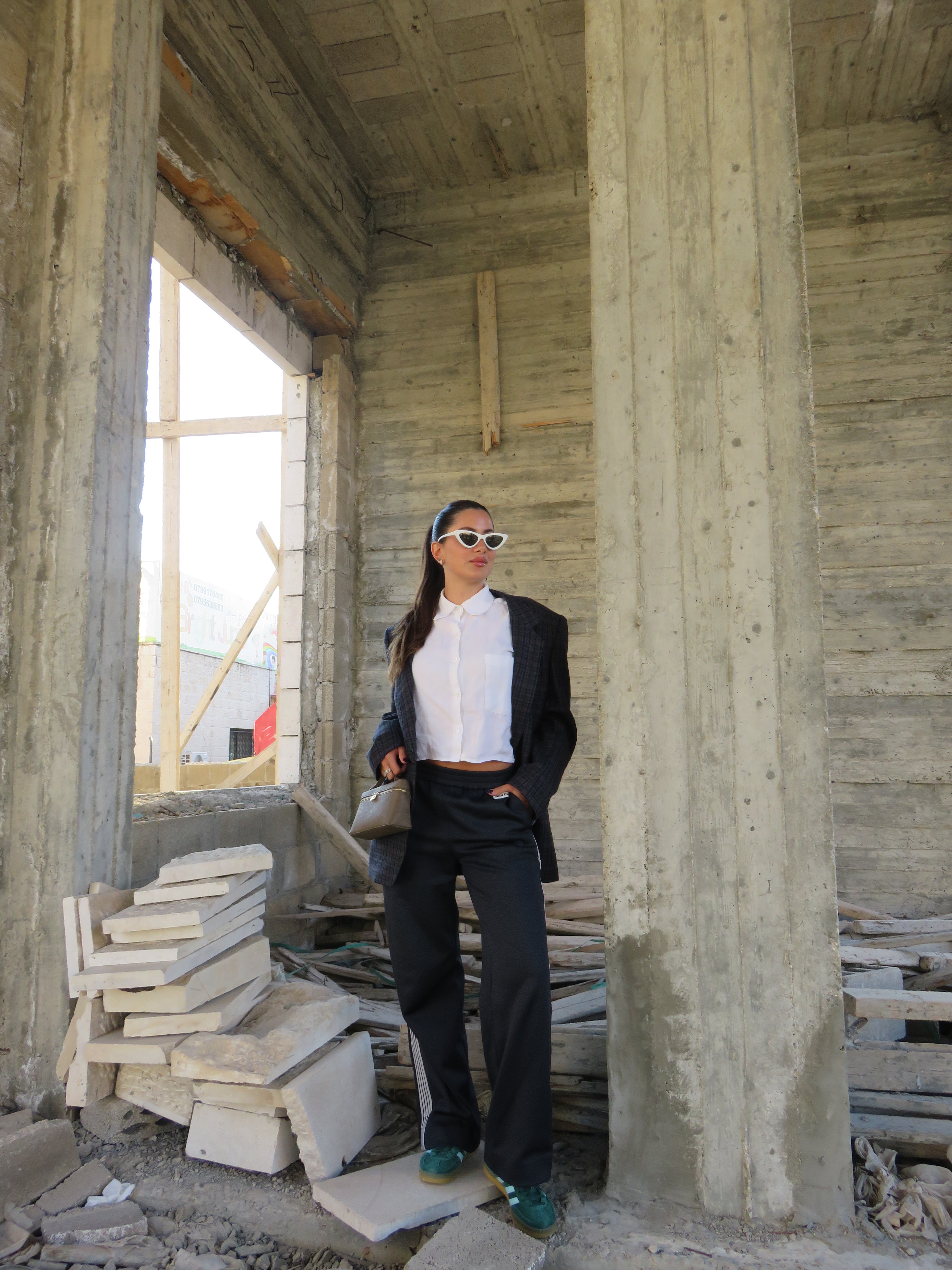 Woman wearing oversized plaid blazer, white cropped shirt, black pants, and white cat-eye sunglasses in unfinished concrete building