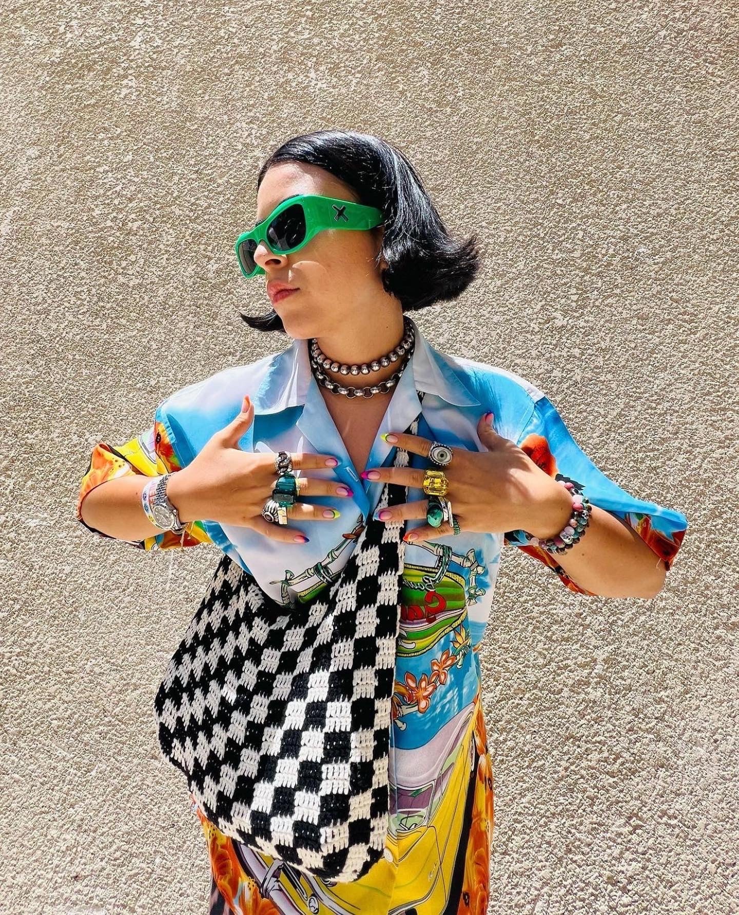Woman in 90s vintage shirt with bold print, green sunglasses, layered rings, and checkered bag