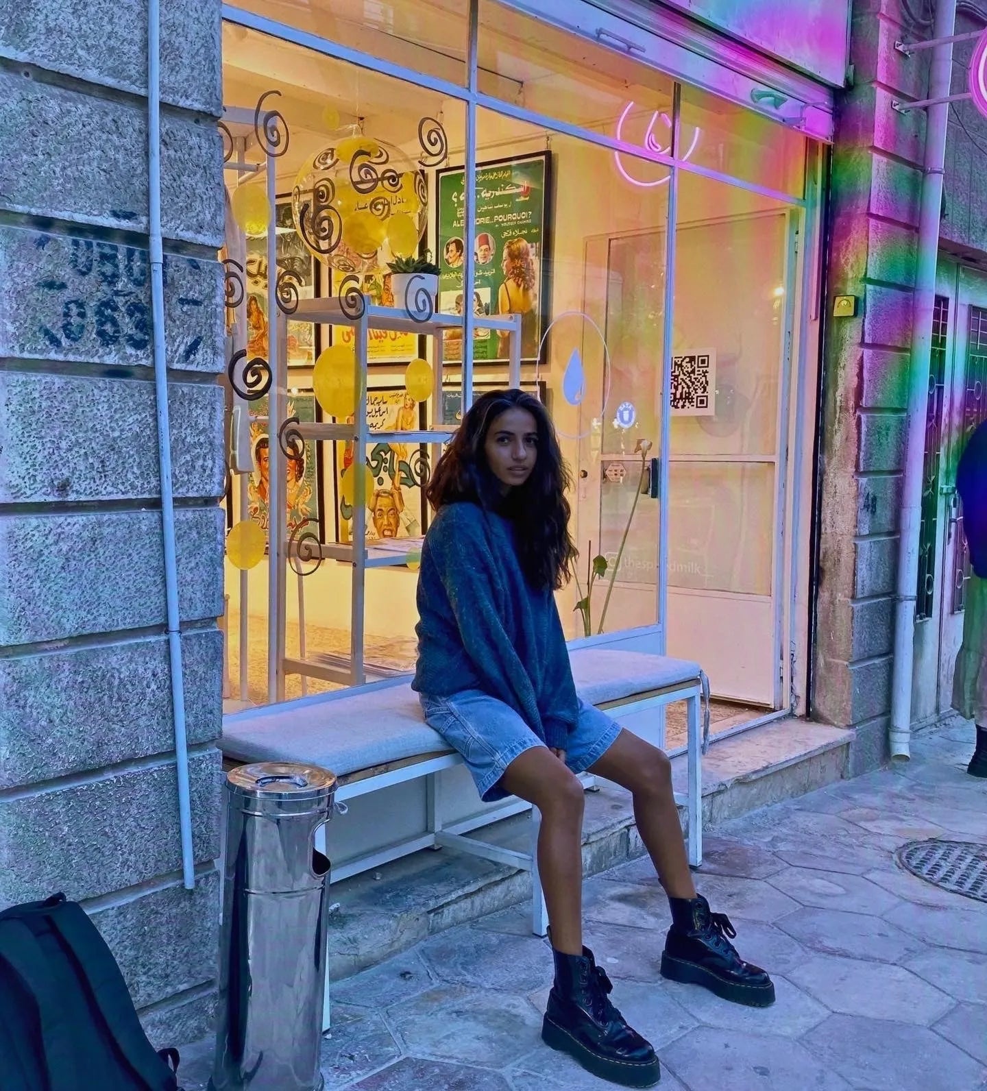Woman in vintage oversized sweater, denim shorts, and chunky boots sitting outside colorful shop